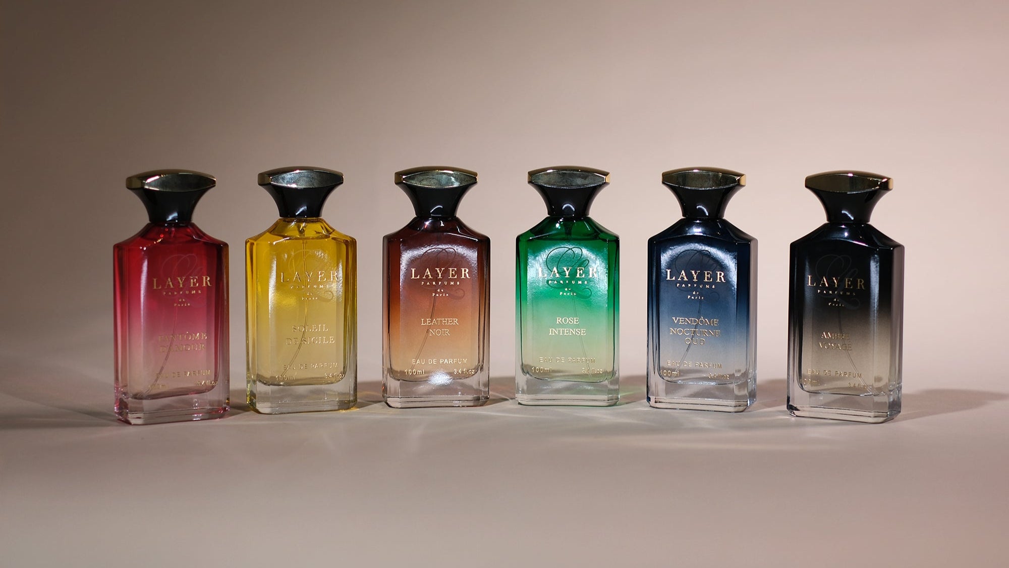 Six rectangular glass perfume bottles in gradient shades of red, yellow, orange, green, blue, and black on a beige surface